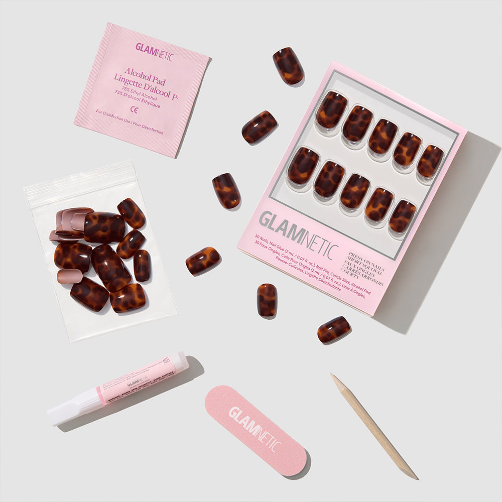A flat lay of Glamnetic's High Stakes press-on nail kit shows short squoval tortoiseshell nails, a pink nail file, alcohol pad, cuticle stick, glue tube, and extra nails arranged on a light gray background. - Product image