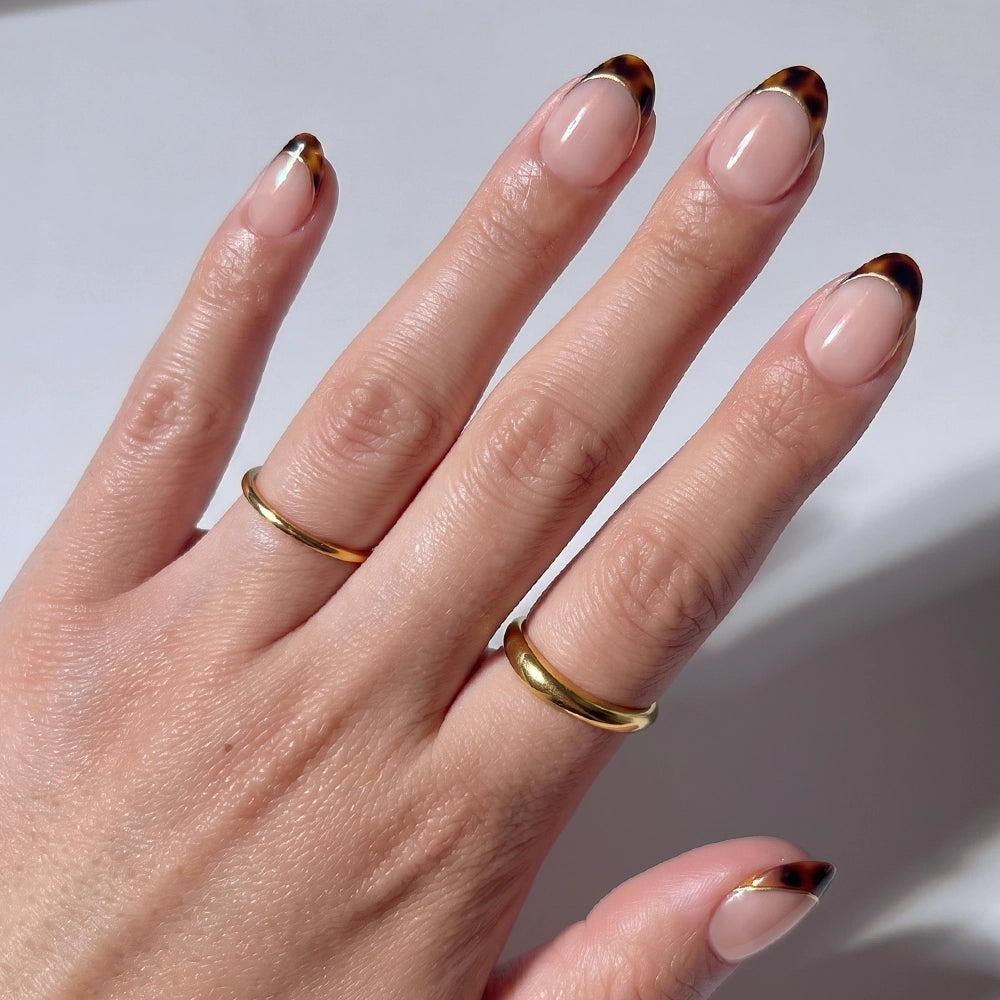 A hand showcases glamnetic’s High Standards almond press-on nails with a brown tortoiseshell French tip, accessorized with two simple gold rings on the middle and ring fingers, set against a neutral background.