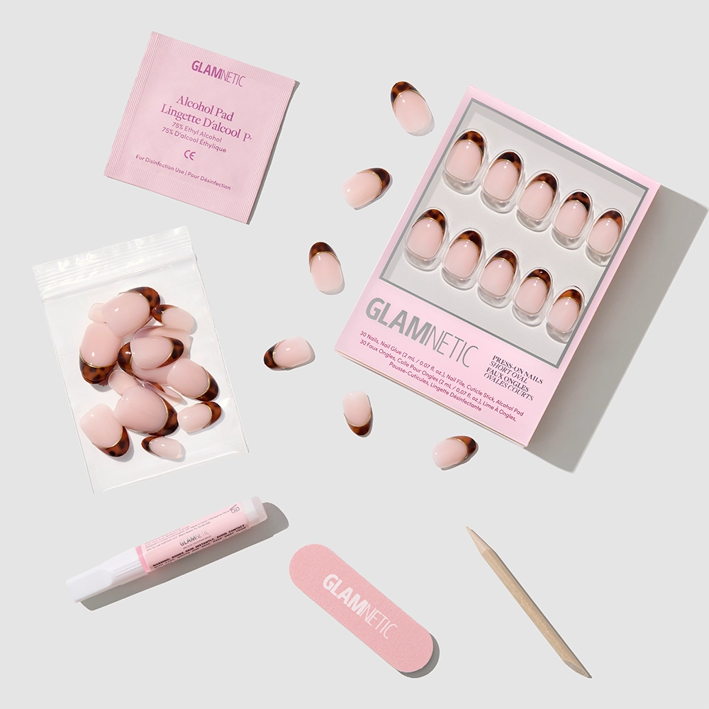 A flat lay of glamnetic’s High Standards pink press-on nails with rose gold tips, short oval shape, mini nail file, cuticle stick, glue, alcohol pad, and extra nails—all neatly arranged on a light background. - Product image