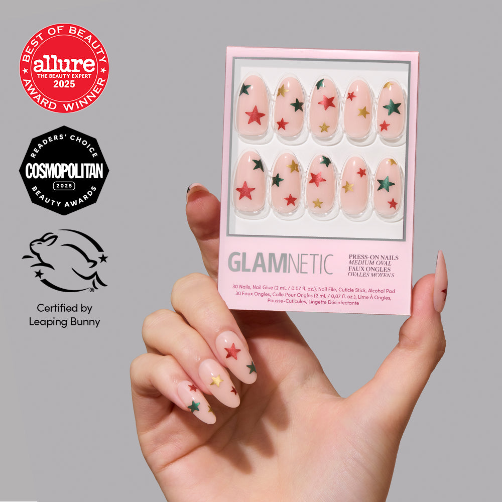 Holiday Wish - A hand holds a Holiday Wish manicure set by glamnetic featuring almond-shaped press-on nails with clear bases and red and green star designs, surrounded by Allure and Cosmopolitan award seals and a Leaping Bunny logo.