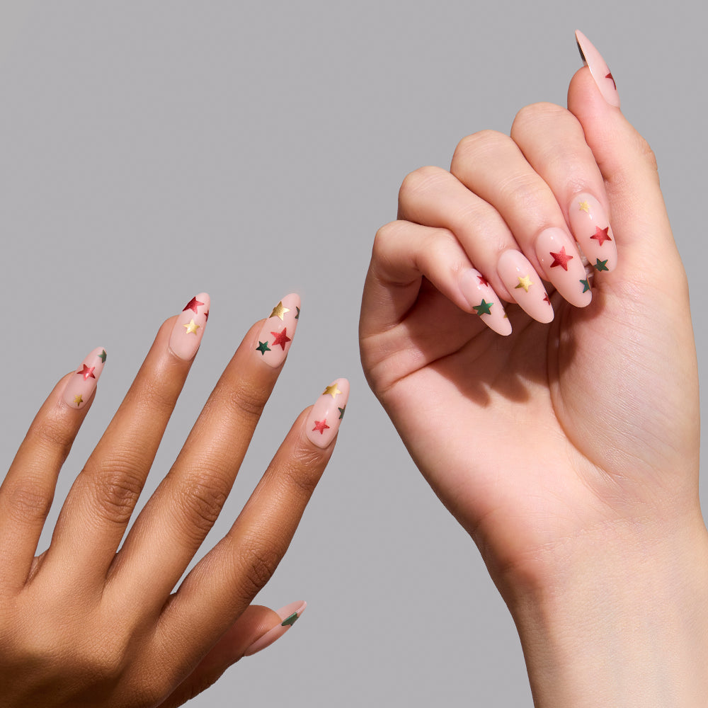 Holiday Wish - Two hands with almond-shaped nails featuring the Holiday Wish set, showing a sheer pink base decorated with small red, gold, and green star decals.