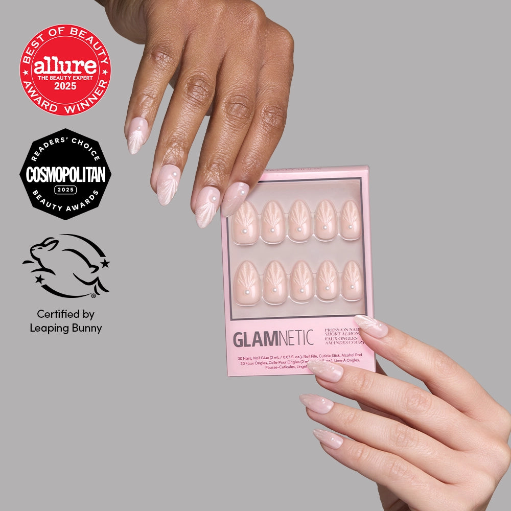 A hand displays short almond-shaped Honeymoon press-on nails by glamnetic with a pale pink base and white swirling leaf designs, next to a bridal nail set box featuring award logos from Allure, Cosmopolitan, and Leaping Bunny.