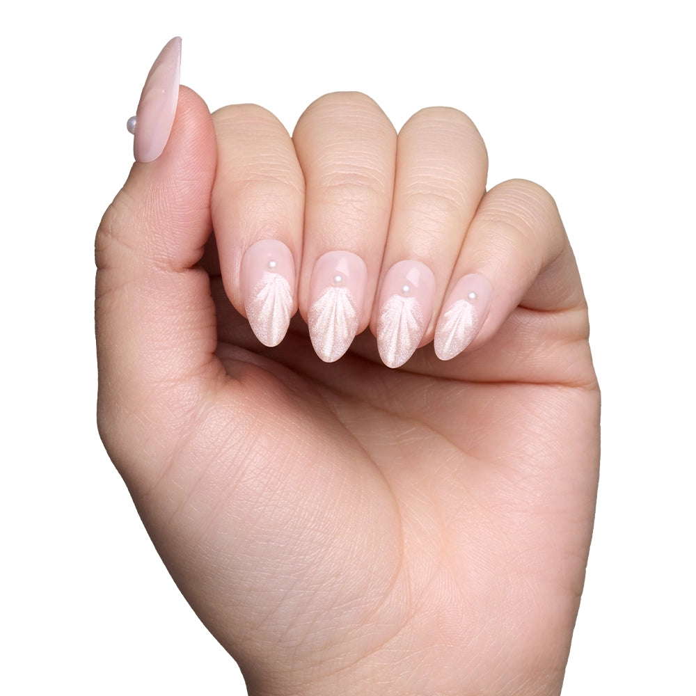 A hand shows off glamnetic’s Honeymoon nails—light pink, almond-shaped press-ons with seashell designs, pearl accents at the base, and delicate 3D swirls—against a white background. - Product image