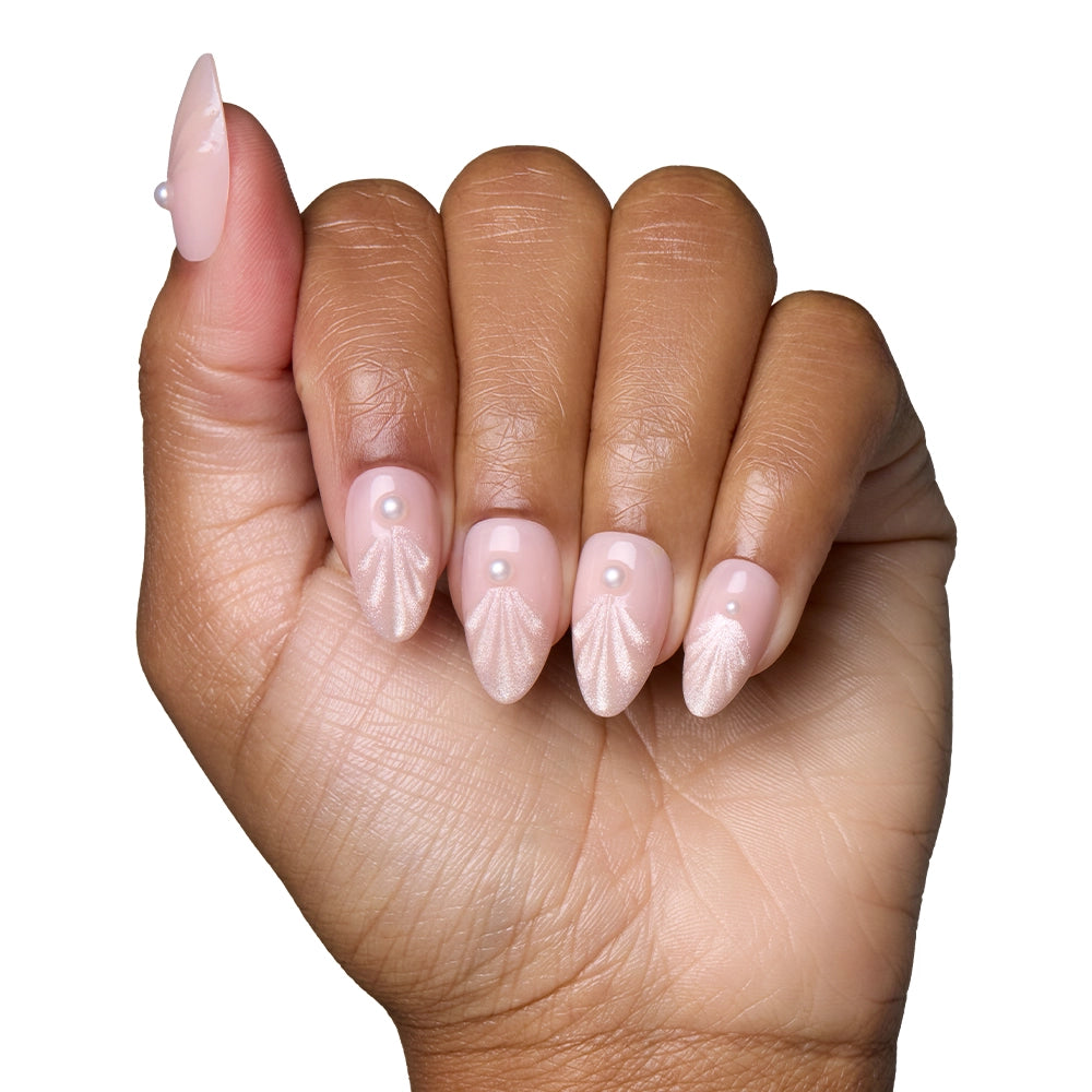 A hand displays the glamnetic Honeymoon Nail Set: almond-shaped nails in soft pink with dainty pearl accents, subtle shimmery seashell designs, and 3D raised swirls for an elegant finish against a white background. - Product image