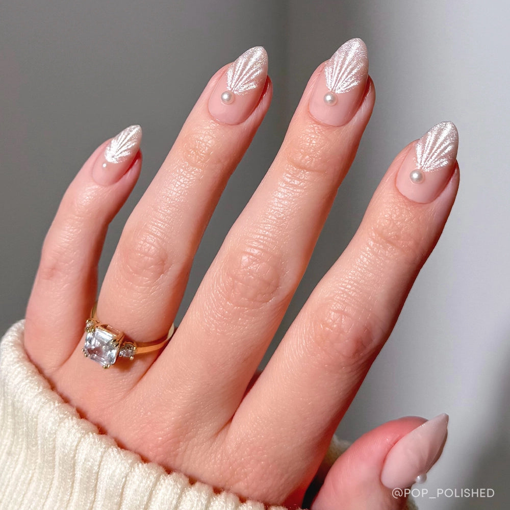 A hand with almond-shaped nails from glamnetics Honeymoon set shows a pale pink base, pearly white leaf designs, and dainty pearl accents. The person wears a gold ring with a large gemstone and a cream-colored sweater. - Product image