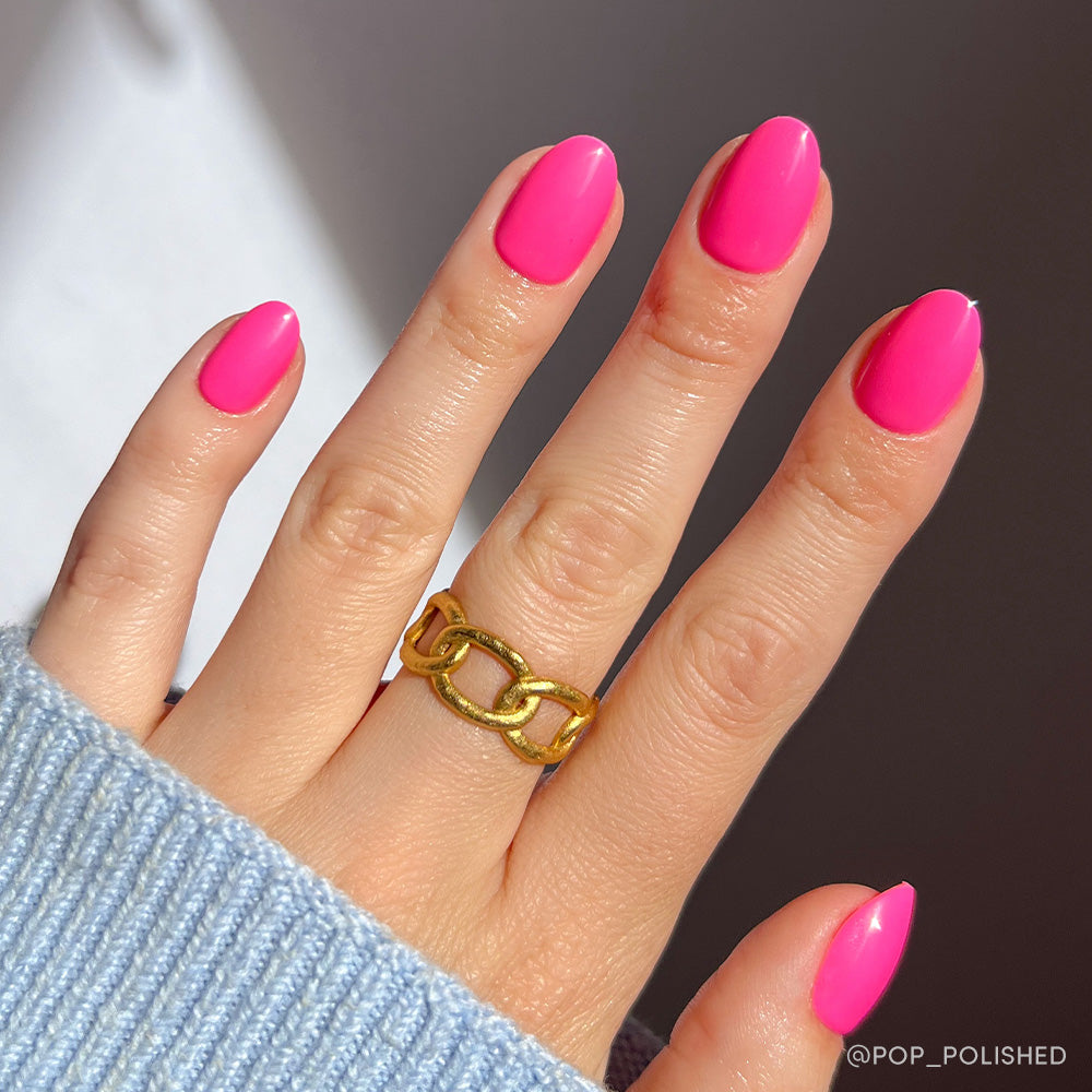 A hand with glamnetic Hot Pink short oval nails and a chunky gold chain ring on the middle finger, partially covered by a light blue knit sleeve.