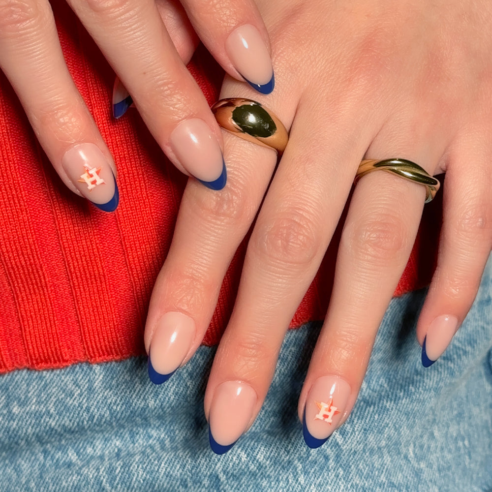 Hands display MLB Press-On Nails by glamnetic—nude nails with blue tips and two accent nails featuring orange Houston Astros H logos. The person accessorizes with gold rings and wears a red sweater paired with blue jeans. - Product image