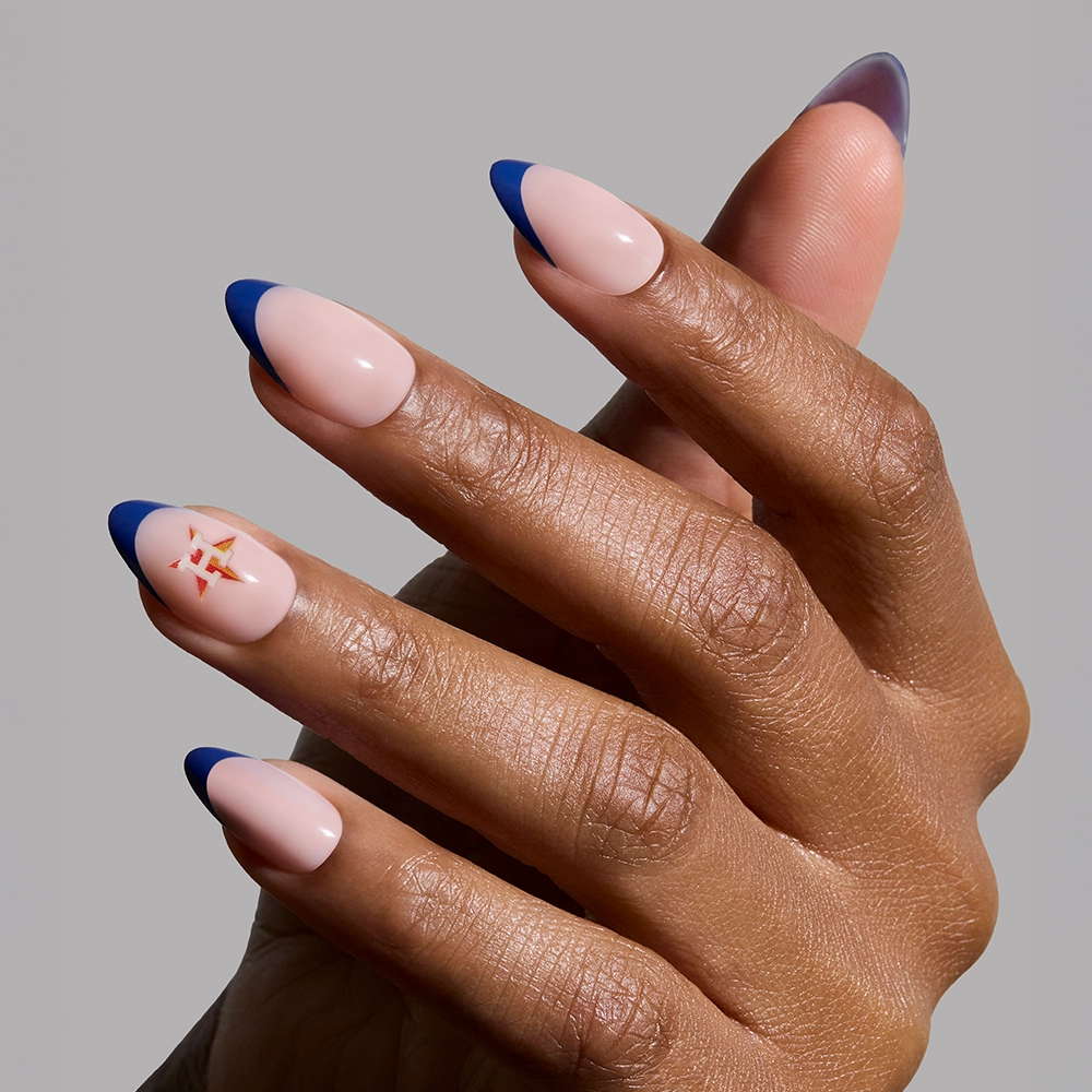 Show off your glamnetic Houston Astros Press-On Nail Set with almond-shaped nude nails and dark blue French tips. The ring finger features a geometric orange and gold accent near the cuticle, all set against a light gray background. - Product image