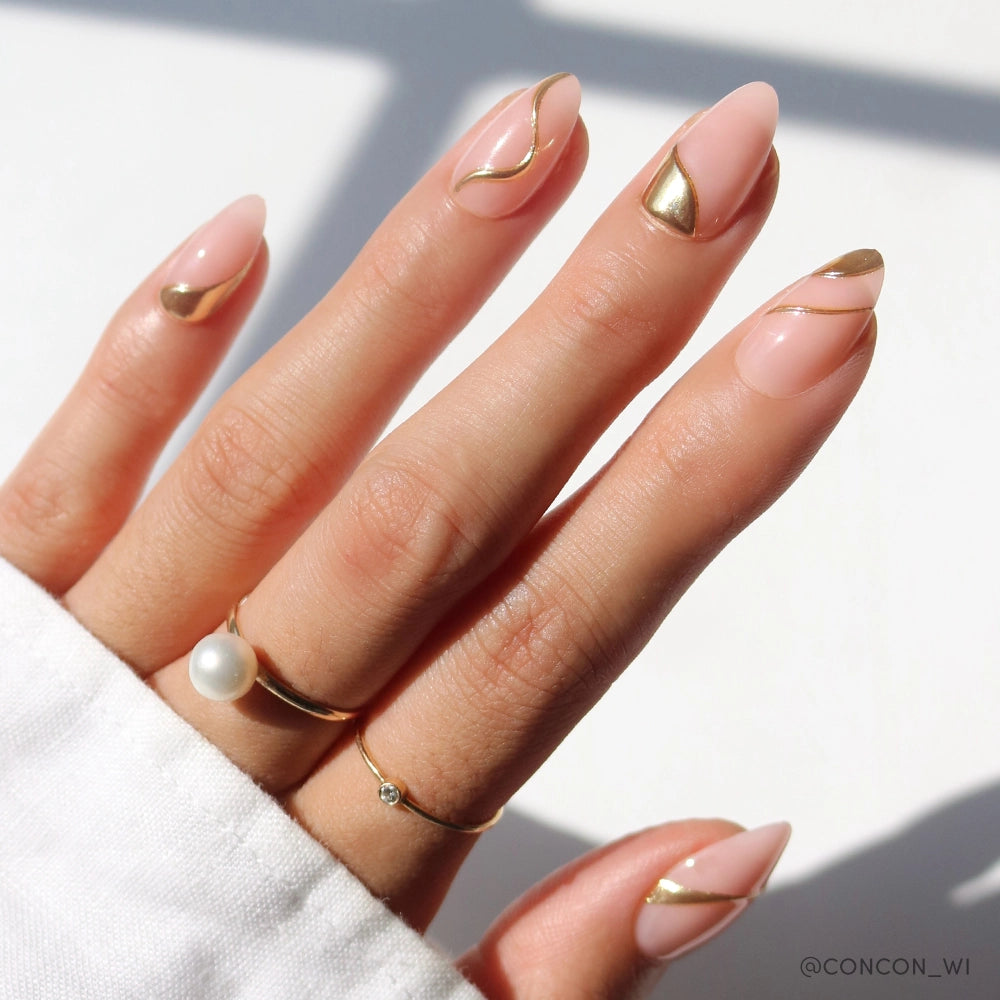 Close-up of a hand with almond-shaped press-on nails in a soft nude-pink base, embellished with asymmetrical gold chrome accents in curved, wave-like patterns. The hand is accessorized with a pearl ring and a dainty diamond ring.

