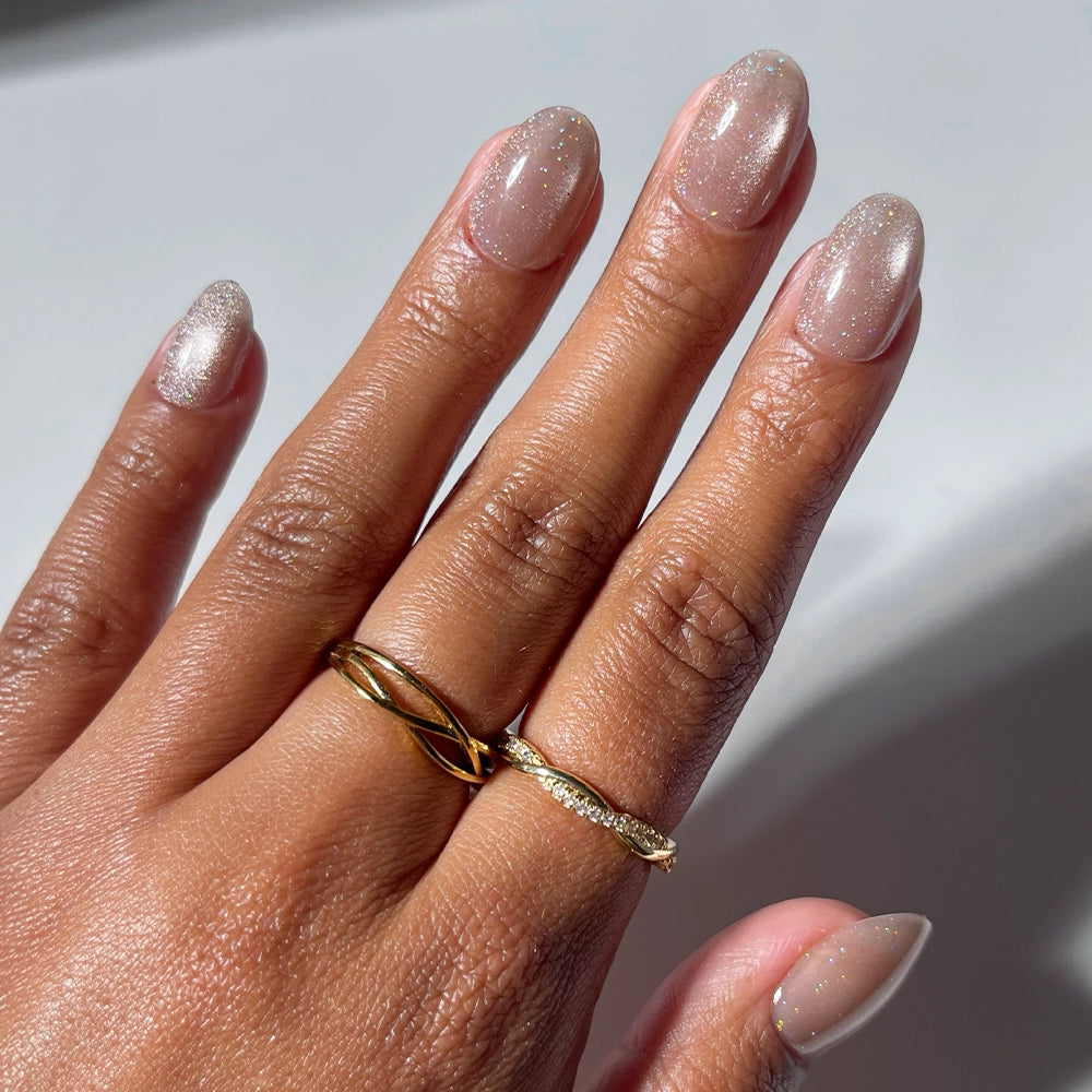 A hand showcases glamnetic’s Iced Latte press-on nails in a glittery nude almond shape, adorned with two gold rings—one featuring a twisted design and the other set with small crystals. This elegant nude mani is perfect for fans of press-on nails, standing out beautifully against the blurred, light-colored background. - Product image