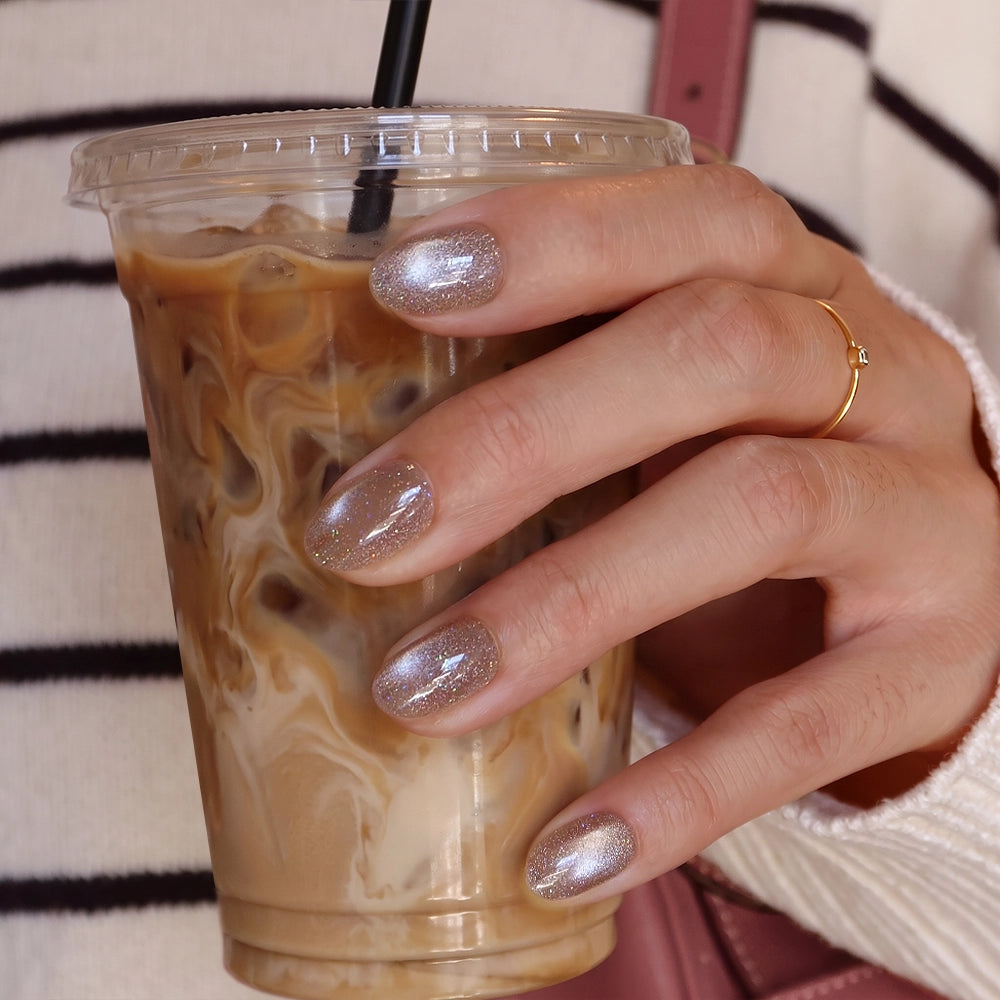 A hand with short oval nails in glamnetic’s shiny silver “Iced Latte” finish holds a plastic cup of iced coffee with a black straw; the person wears a thin gold ring and a striped sweater. - Product image