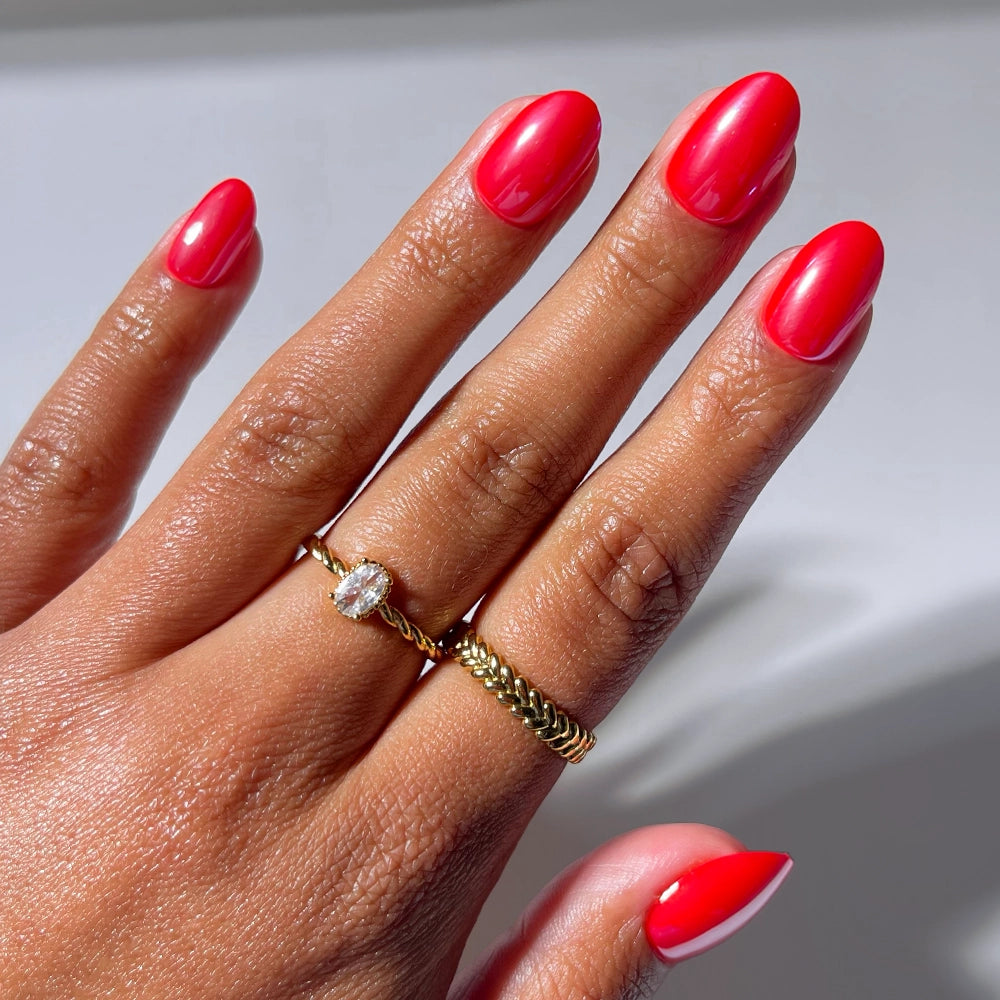 A hand with glossy, bright pink "Icon Red" press-on nails by glamnetic wears two gold rings—one plain band and one with a small oval gemstone—set against a neutral background.