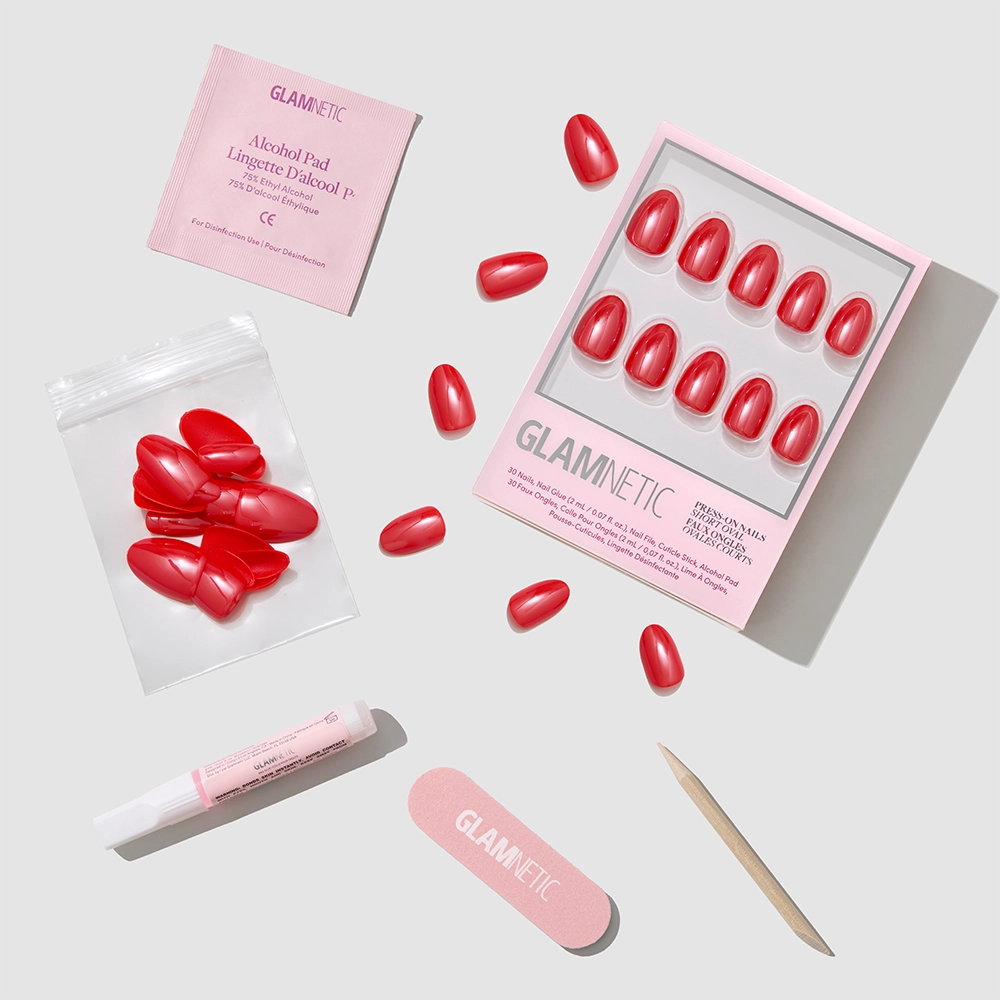 Icon Red by glamnetic: red short oval press-on nails with an alcohol pad, nail glue, file, wooden stick, and extra nails—all neatly arranged on a light gray background.
