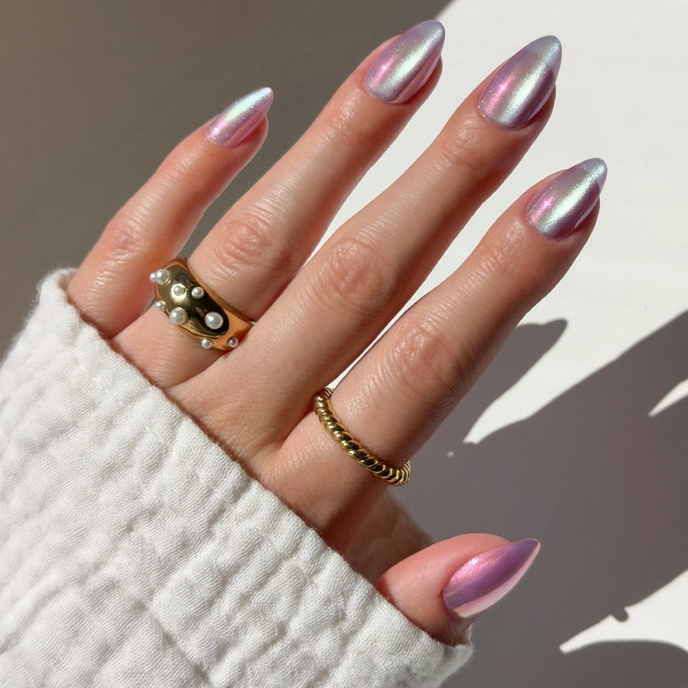 A hand with long, pearlescent pink and purple glamnetic Iconic Oyster press-on nails is shown. The person wears a white textured sleeve and three gold rings, one with small pearls, against a soft neutral background.