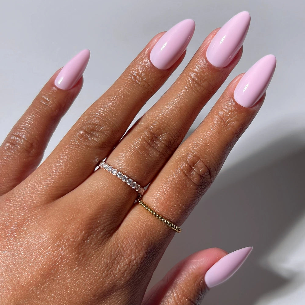 A hand with medium-brown skin shows off long, almond-shaped nails painted in glamnetic’s Iconic Pink. On the middle and ring fingers are a thin diamond ring and a simple gold band.