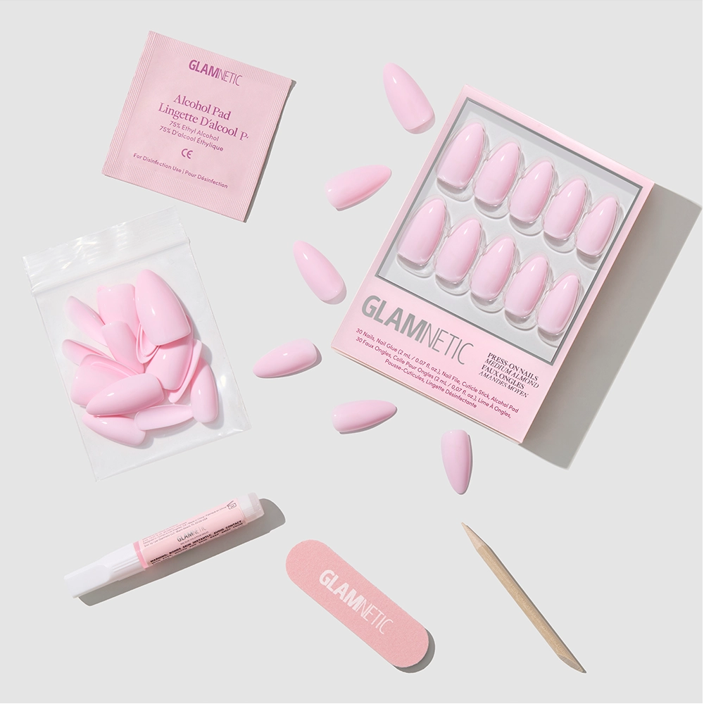 The glamnetic Iconic Pink set includes almond-shaped pink press-on nails with a glossy finish, a nail file, wooden cuticle stick, glue, extra nails in a bag, and an alcohol pad—all neatly arranged on a light surface.