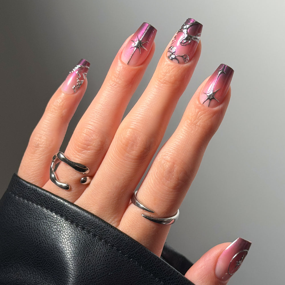 A close-up of a hand showcases glamnetic’s Immortal Kiss press-on nails, featuring long, coffin-shaped glossy ombré tips with a pink-to-purple gradient and adorned with silver chrome star and molten-style accents. The hand is accessorized with two sculptural silver rings and the sleeve of a black leather jacket.