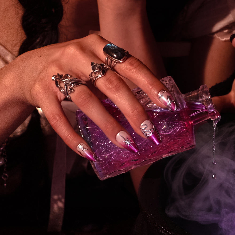 A hand with long, glossy ombre nails and multiple silver rings holds a glass bottle of glamnetic’s Immortal Kiss, its purple liquid spilling out and creating a mystical mist. - Product image
