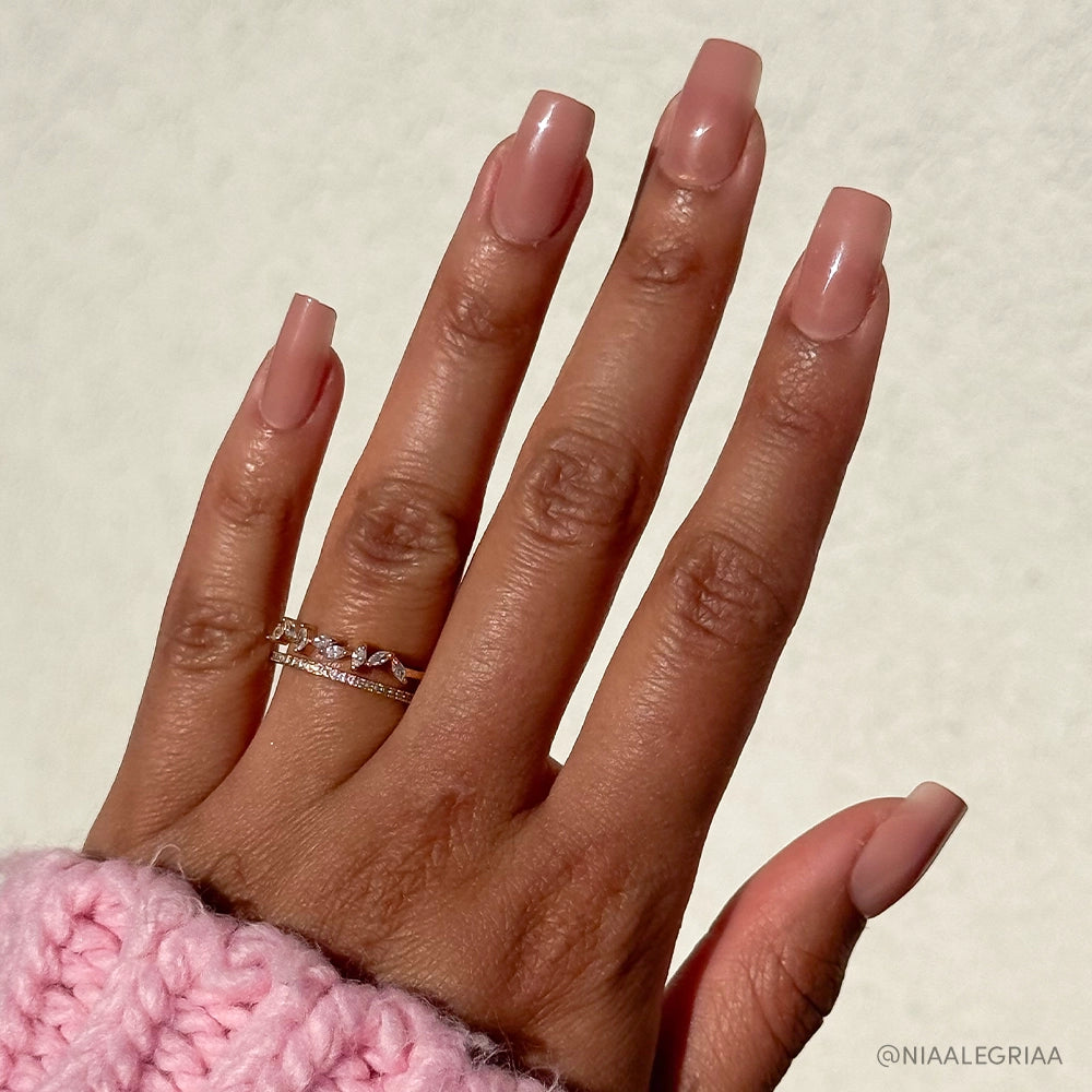 A hand with glamnetic’s “In The Nude” square-shaped, nude-pink ombré nails appears against a light background. Two gold rings adorn the fingers, while a pink knitted sleeve covers the wrist. This vegan, cruelty-free manicure is minimalist.