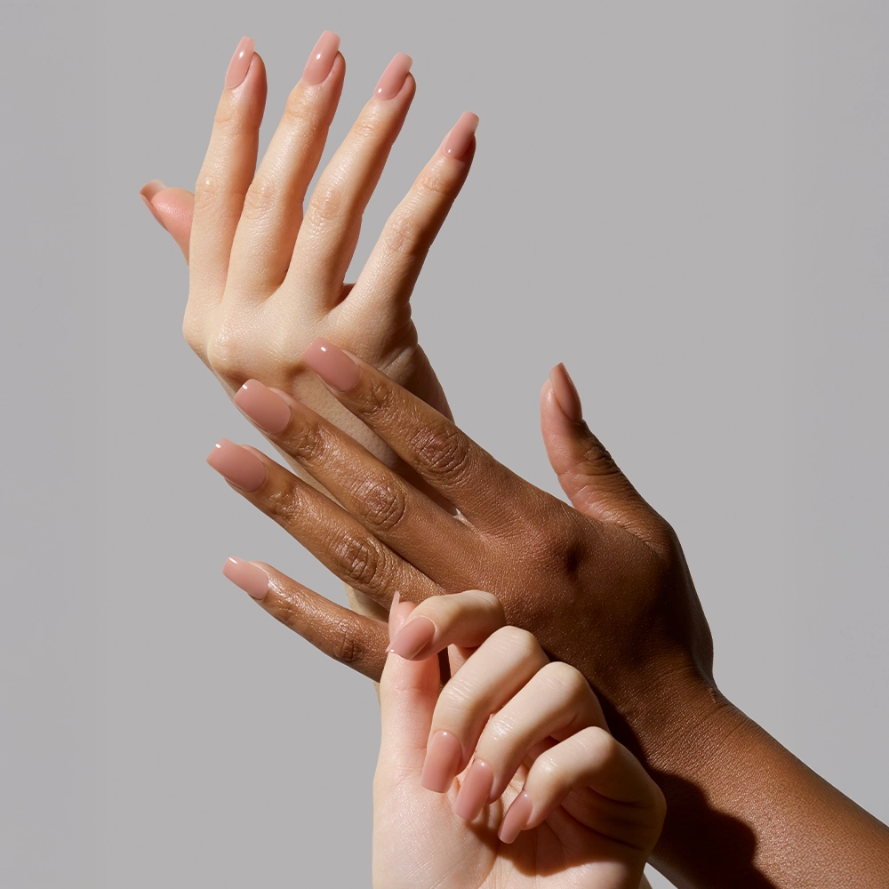 Three hands with varying skin tones display neatly manicured nails painted in glamnetics In The Nude, a nude-pink ombré polish, gracefully posed together against a plain light gray background. - Product image