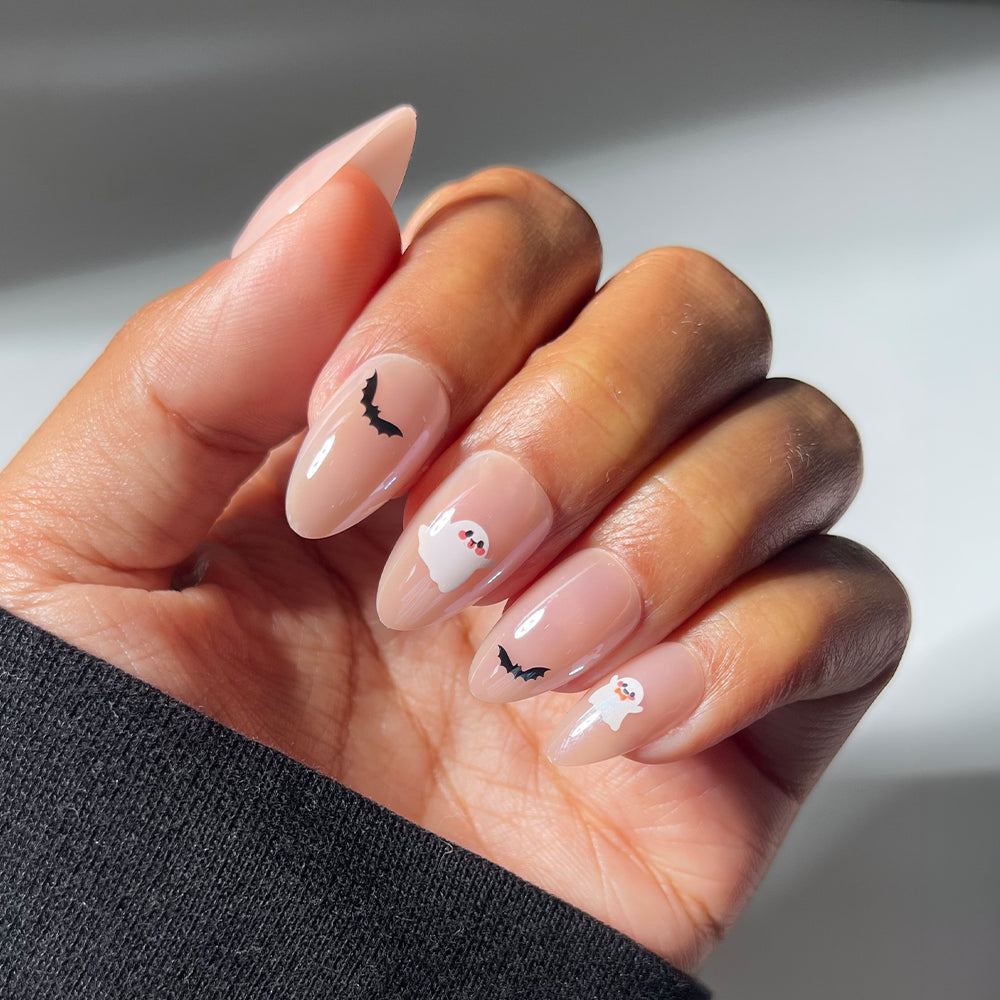 A hand with short almond nails in glamnetic’s “In The Spirit” style, featuring a glazed finish and decorated with Halloween nail art—small black bats and white ghosts on the middle and ring fingers. The person is wearing a black sleeve.