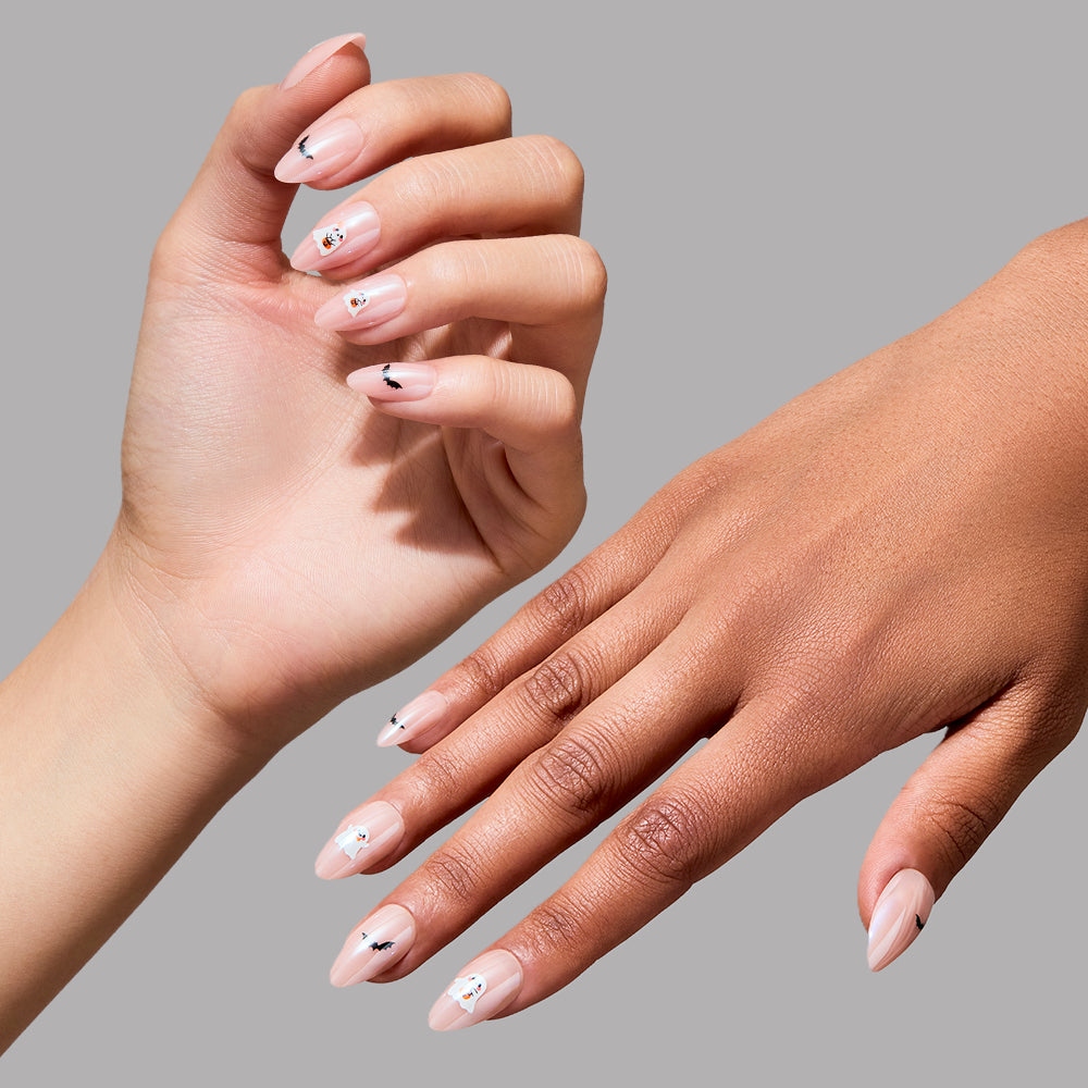 Two hands with short almond nails painted in “In The Spirit” by glamnetic—a sheer, light pink polish with a delicate glazed finish. Each nail is adorned with adorable Halloween-themed stickers, including tiny white ghosts and black bats, set against a simple gray background. - Product image