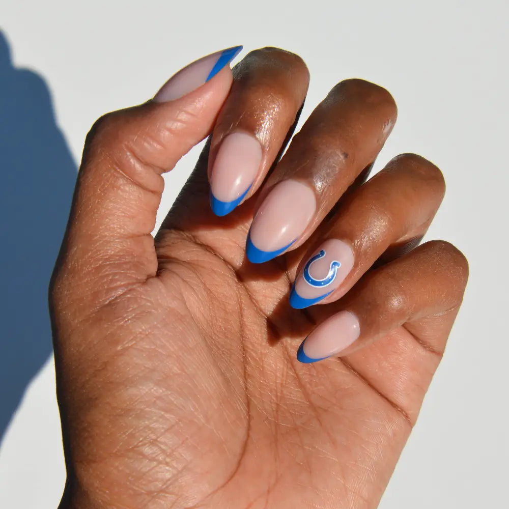A hand adorned with glamnetic’s NFL Press-On Nails, featuring almond-shaped tips in nude polish, striking blue tips, and a blue swirl accent on the ring finger, displayed against a plain light background. - Product image