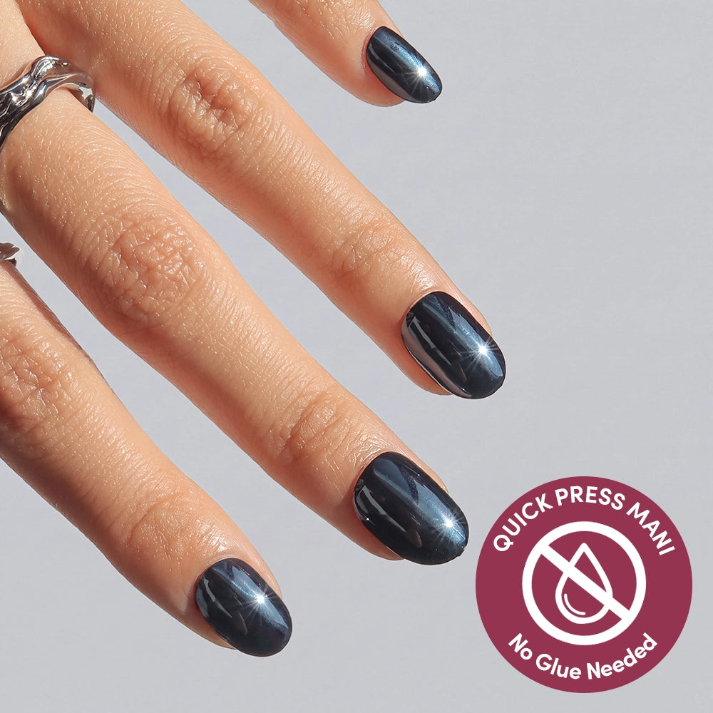 A hand with shiny dark blue Ink Spell press-on nails by glamnetic is shown against a light background. A circular badge reads QUICK PRESS MANI No Glue Needed, with a crossed-out glue bottle icon, emphasizing the easy-to-apply, no-glue design of these nails.
