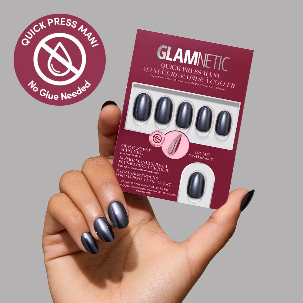 A hand with shiny black press-on nails holds a glamnetic Ink Spell Quick Press Mani package, displaying eight extra stick-on nails and text stating No Glue Needed against a gray background. - Product image