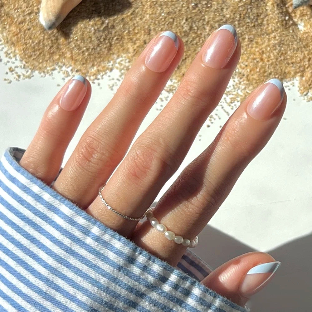 A hand with short oval nails, styled with glamnetic’s Into Blue press-ons in a blue and white French manicure, wearing two delicate rings, rests on a surface with sand and a seashell in the background. The sleeve is blue and white striped.