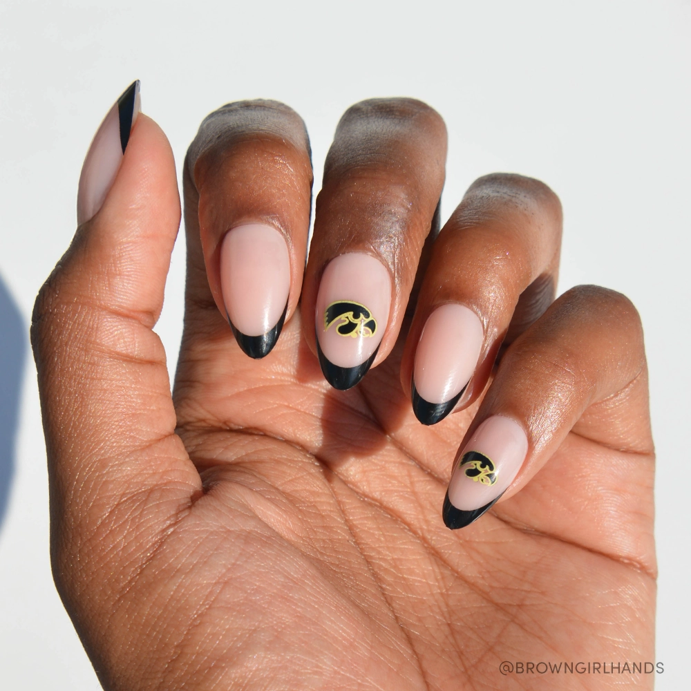 A hand wears the glamnetic Iowa Hawkeyes Press-On Nail Set, showing almond-shaped nails with a nude base, black French tips, and gold leaf accents on two nails, photographed against a white background.