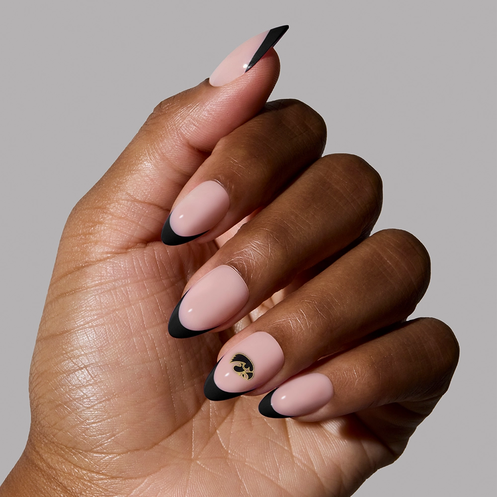 A hand displays the glamnetic Iowa Hawkeyes Press-On Nail Set in nude with black French tips, featuring a gold and black accent design on one nail, against a plain gray background. - Product image