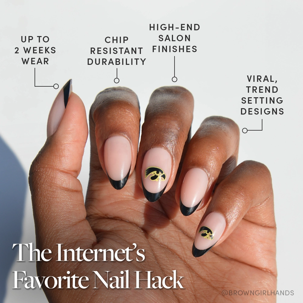 A hand with long, almond-shaped nails featuring nude polish, black French tips, and gold accents from the officially licensed Iowa Hawkeyes Press-On Nail Set by glamnetic. Enjoy 2 weeks wear, chip resistance, salon finishes, and trendy designs. - Product image