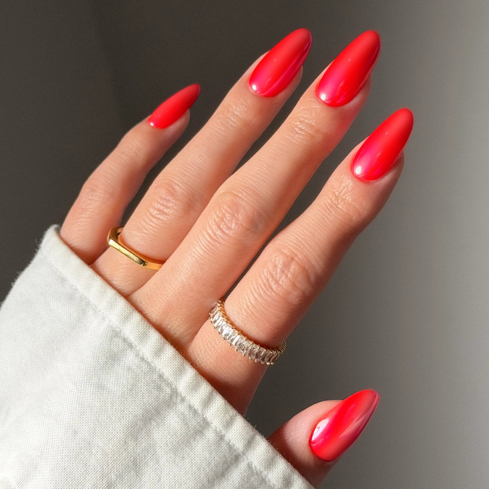 A hand with long, glossy It-Girl Red press-on nails by glamnetic in a medium oval shape wears gold and diamond-studded rings. The person is dressed in a light-colored, long-sleeve shirt.