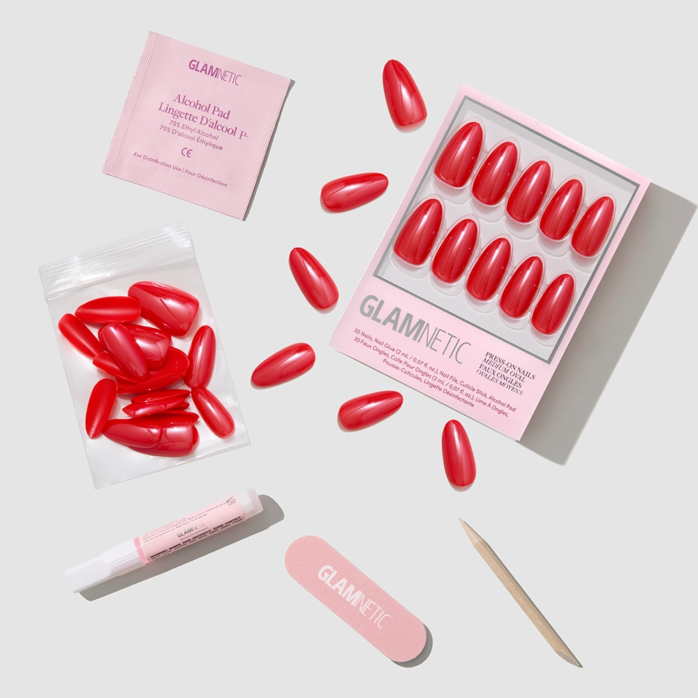 A set of red cruelty-free false nails, glue, alcohol pad, nail file, wooden cuticle stick, and extra nails are arranged neatly on a light gray background. The packaging reads "It-Girl Red" by glamnetic.