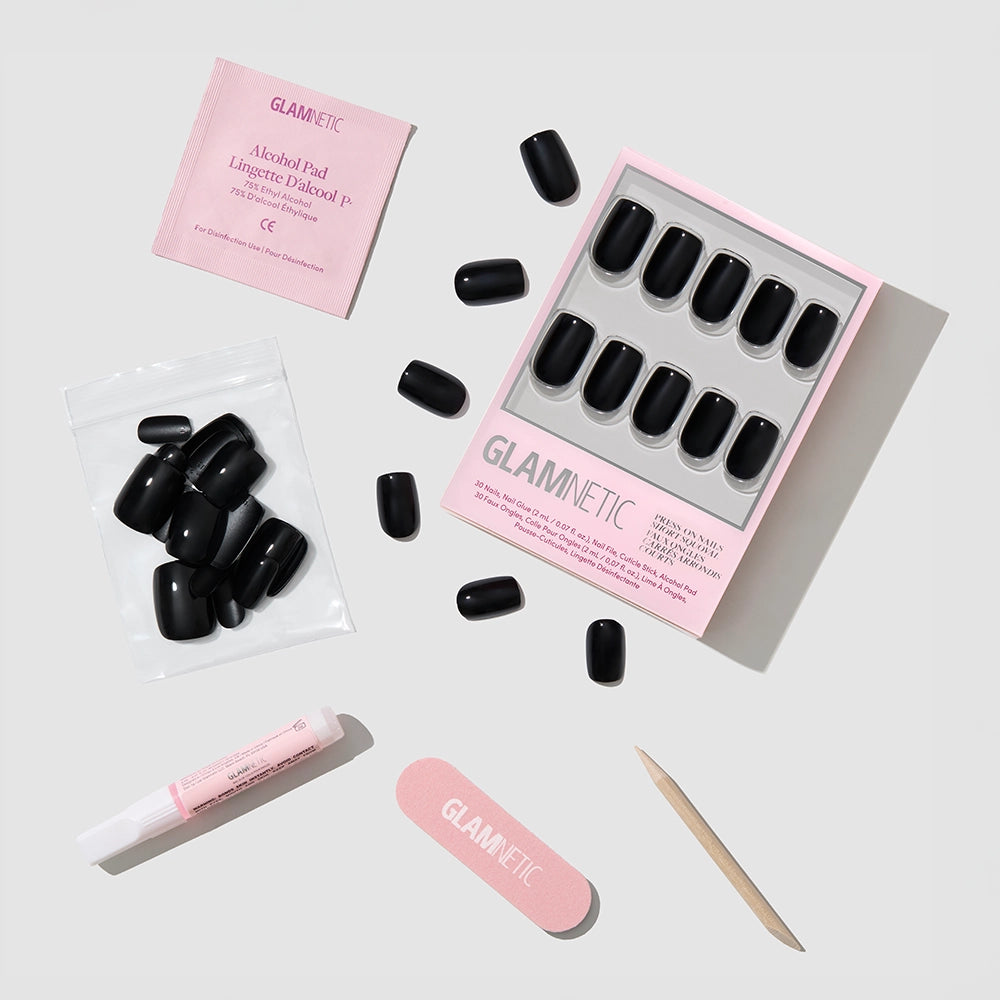 A flat lay of the glamnetic Jet Black set displays glossy squoval nails, nail glue, a wooden cuticle stick, pink file, alcohol pad, and clear bag—all you need for reusable vegan nails—on a light gray background. - Product image