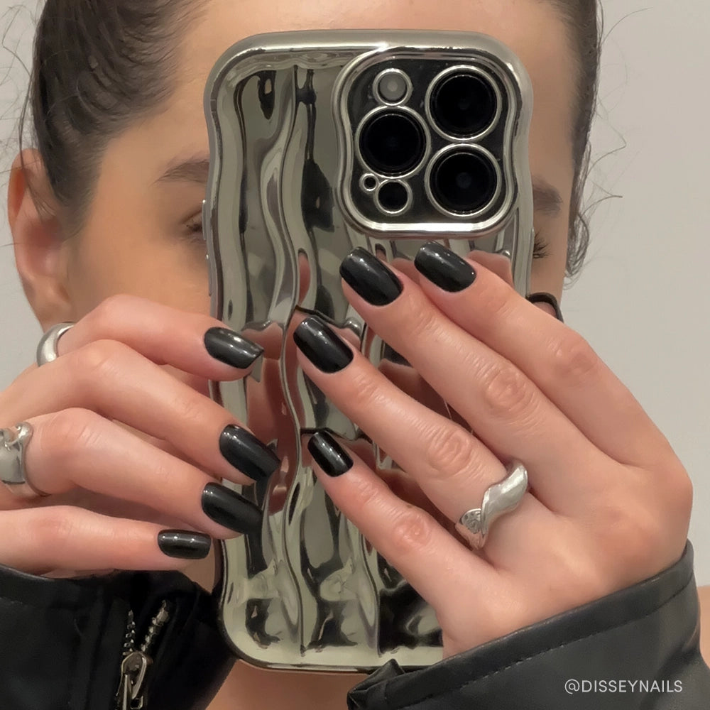 A person with glossy Jet Black squoval nails by glamnetic holds a wavy metallic silver phone case, partially covering their face. They wear silver rings and a dark jacket, capturing the shot as a mirror selfie. - Product image