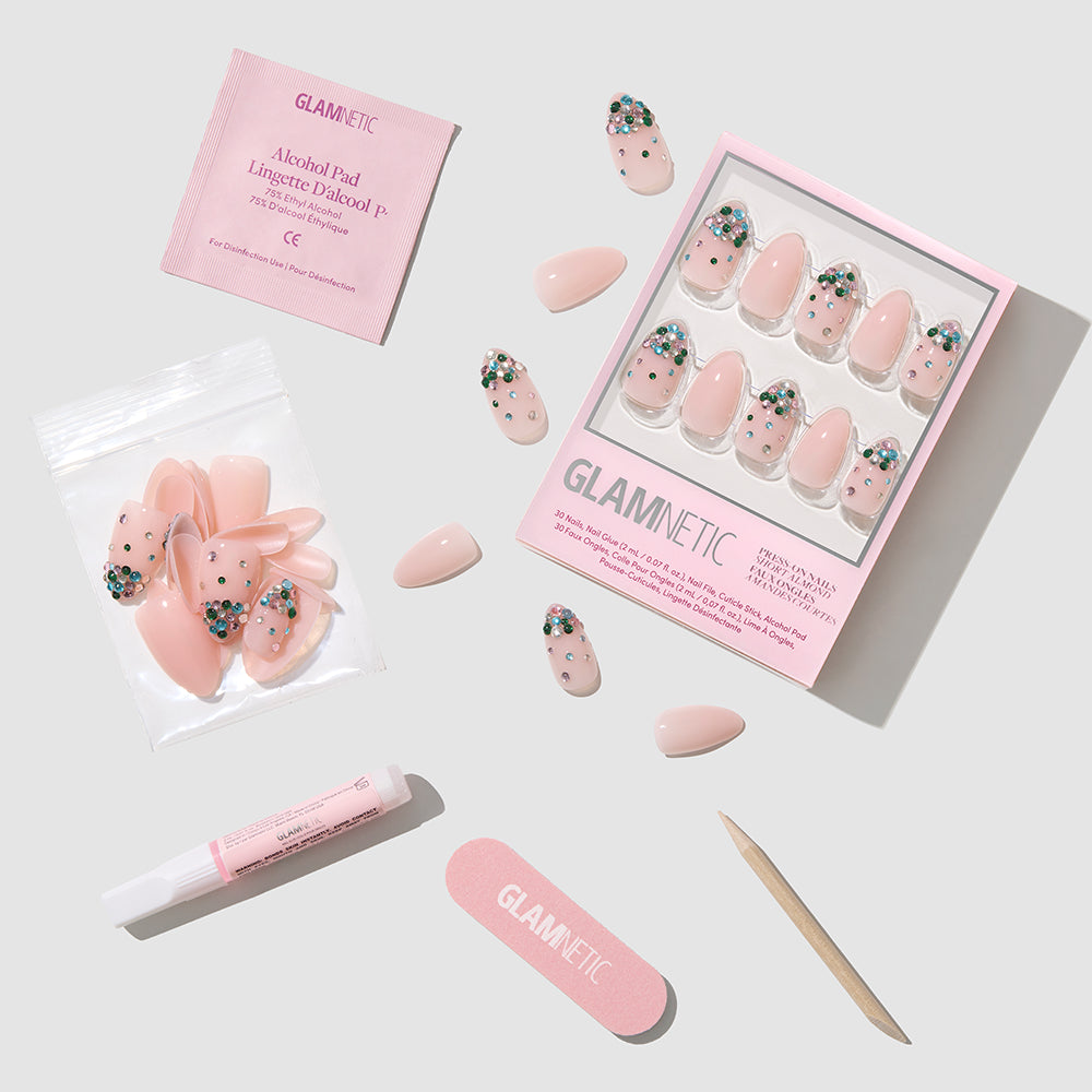 Jewel Thief - Glamnetic Jewel Thief light pink almond press-on nails with green and clear gem accents, shown with a box, extra nails, glue, an alcohol pad, a nail file, and a wooden cuticle stick.