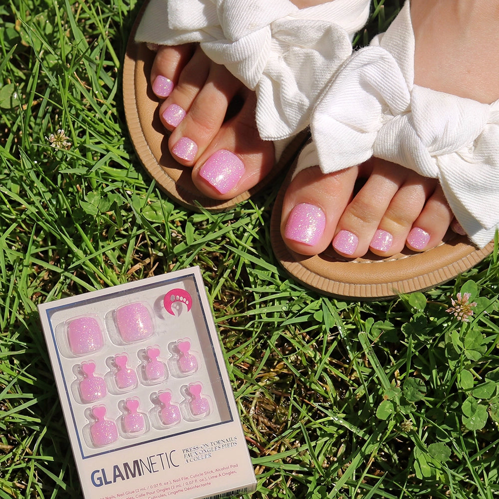 A person in sandals with big white bows displays sparkly pink glamnetic Juicy Toenail press-ons while standing on green grass beside a GLAMNETIC glitter press-on toenail package.