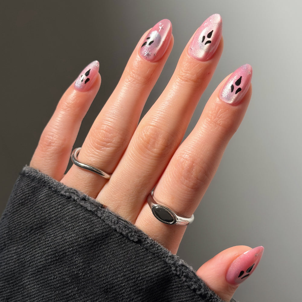 A hand showcases glamnetic’s “Just A Ghoul” short almond press-on nails in a light pink, glittery finish with black ghost face designs. Two silver rings accent the mani, while a dark, frayed-edge sleeve partially covers the hand.