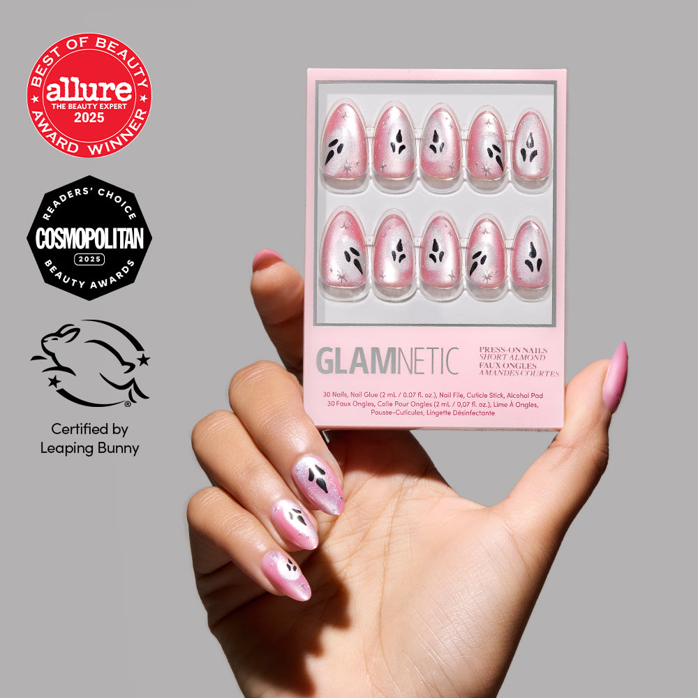 A hand with a short almond mani featuring pink and black floral press-on nails holds a box of glamnetic Just A Ghoul nails. Award logos from Allure, Cosmopolitan, and Leaping Bunny are displayed on a gray background. - Product image