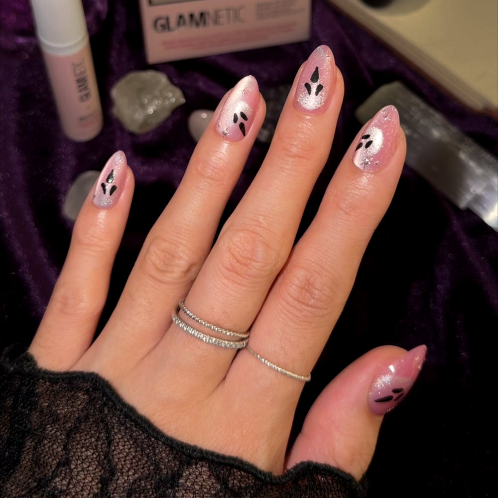 A hand with shiny pink nails in the glamnetic “Just A Ghoul” short almond manicure, featuring black abstract designs and two thin silver rings, rests on a purple velvet surface surrounded by crystals and beauty products. - Product image