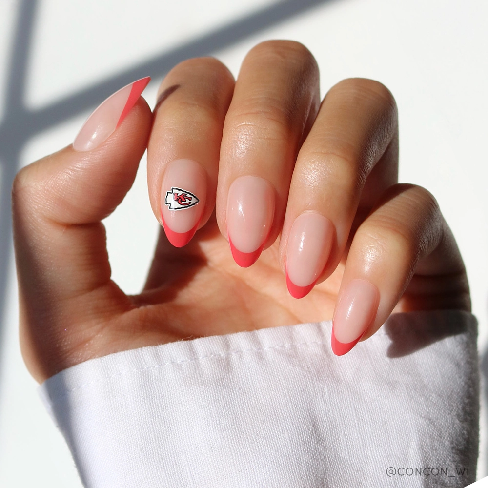A hand with almond-shaped nails wearing the glamnetic Kansas City Chiefs Press-On Nail Set—light pink base, red French tips, and Chiefs art on the ring finger. The person is dressed in a white long-sleeve shirt.