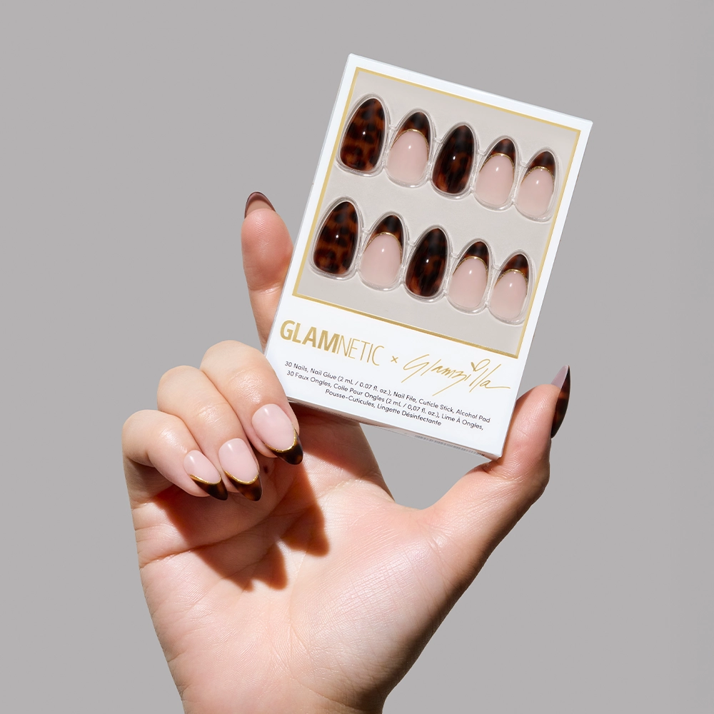 A hand with elegant almond-shaped nails showcasing alternating designs: glossy tortoiseshell full coverage and nude base with tortoiseshell French tips outlined in gold. The person is wearing minimalist gold rings and a cozy white sweater.
