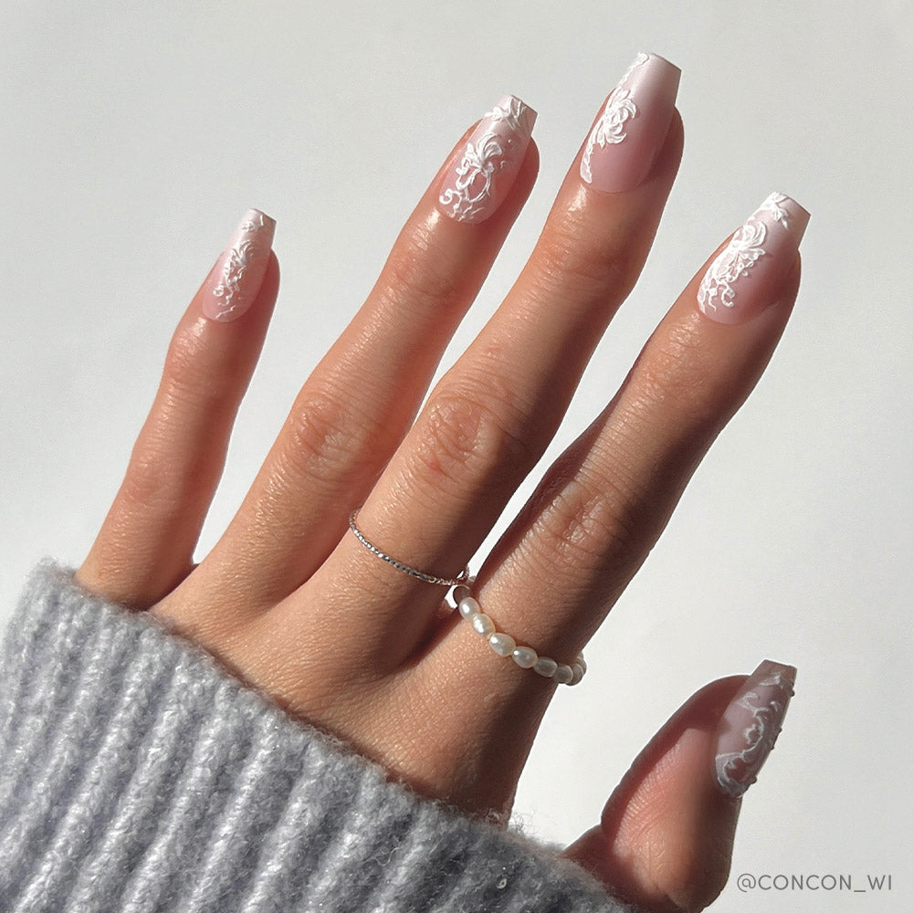 A hand with long, coffin-shaped Lace Blush nails by glamnetic, showcasing intricate white floral and lace designs on a pale pink base. The person wears two delicate rings and a gray knit sleeve partially covers their hand.