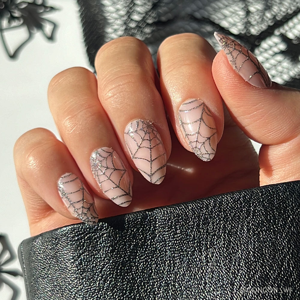 A hand with long, almond-shaped glamnetic Lady Web nails in nude, each featuring a black sparkly spiderweb design, holds textured black fabric—perfect for chic Halloween style. - Product image