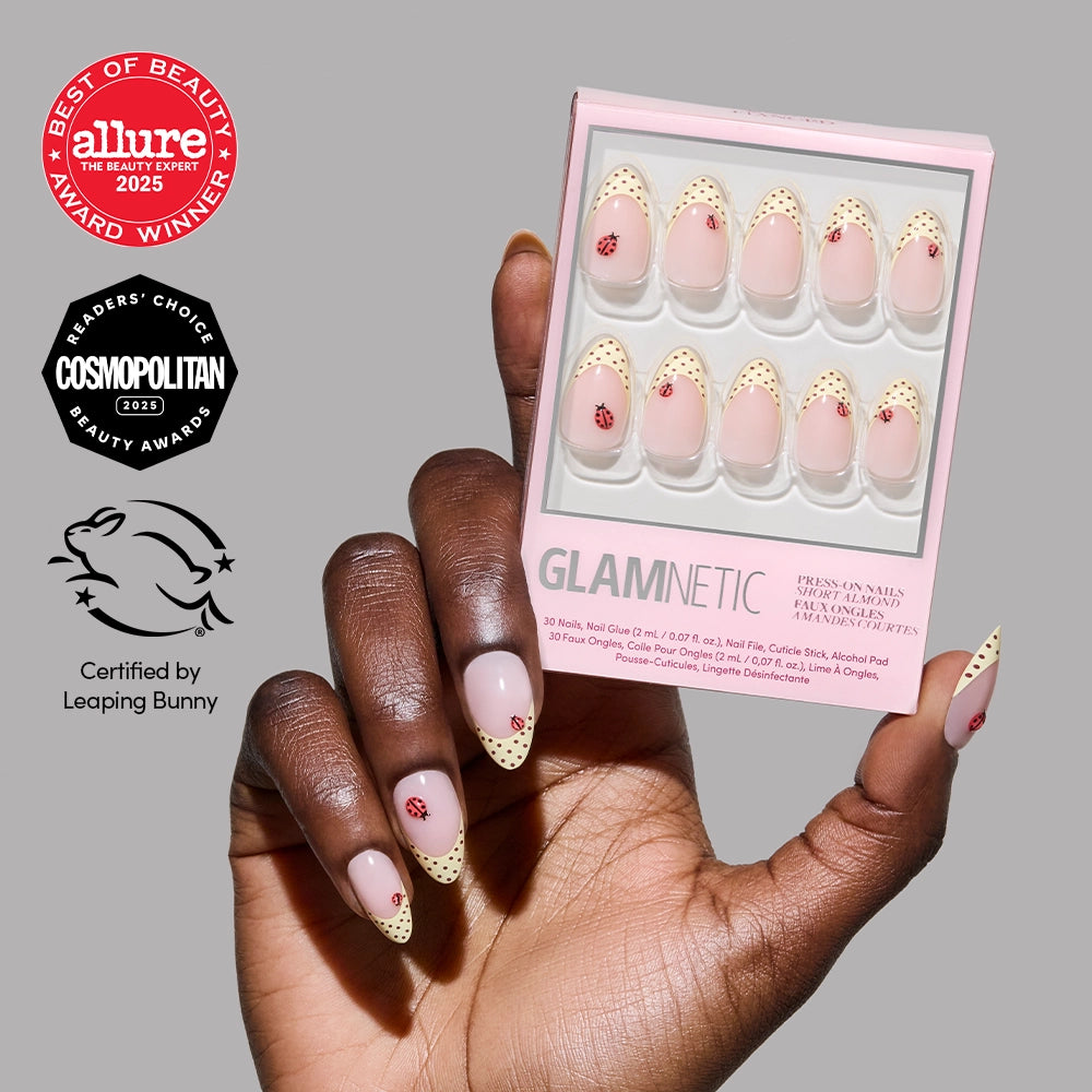 A hand with short almond glamnetic Lady Luck press-on nails—pale base, red cherry art, pale yellow tips with brown dots—holds a matching box. Award badges and Leaping Bunny cruelty-free certification are shown, perfect for summer manicures.