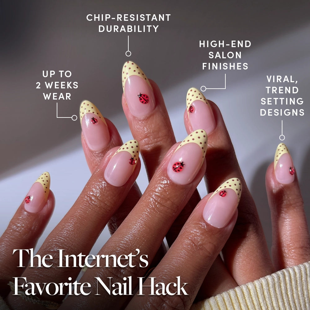 Close-up of hands with short almond nails in Lady Luck by glamnetic, featuring yellow French tips, playful polka dots, and cute ladybug art. Text highlights chip-resistant durability and salon-quality, long-lasting press-on nails. - Product image