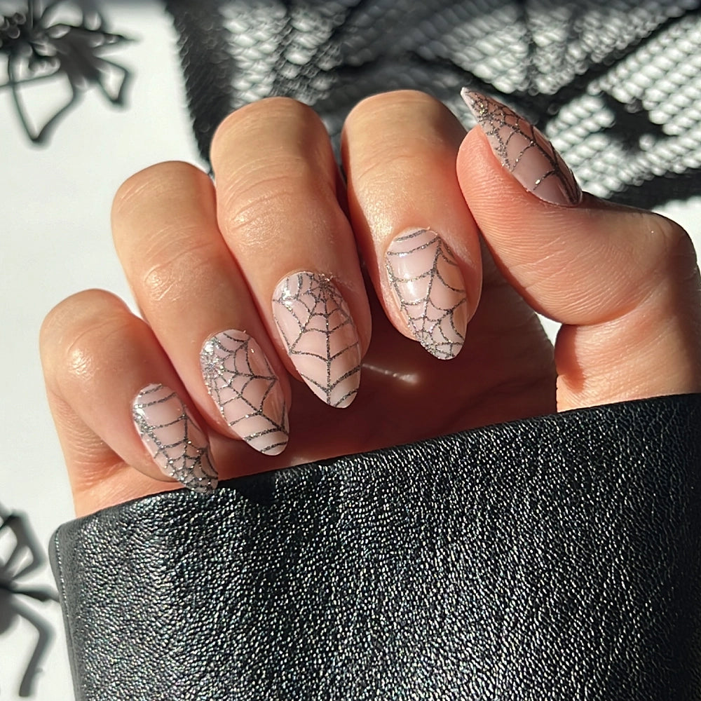 A hand with long, almond-shaped nails showcases the “Lady Web” press-on nails by glamnetic—featuring a sheer base and glittery black spiderweb design—held against a piece of textured black fabric, making it the perfect Halloween mani for glamnetic fans.