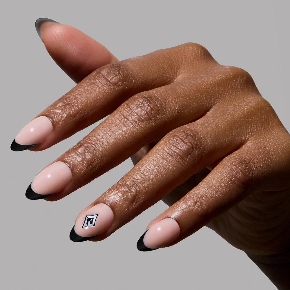A hand with almond-shaped glamnetic Las Vegas Aces Press-On Nails in a nude shade with black tips and a small square geometric accent on the ring finger, all showcased against a plain gray background.